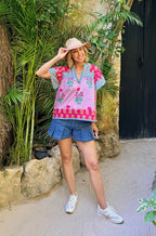Woman in colorful embroidered top and denim shorts standing outdoors with plants and a stone wall in the background.