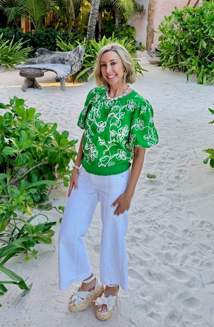 Woman in a green floral blouse and white pants standing on a sandy path with greenery around.