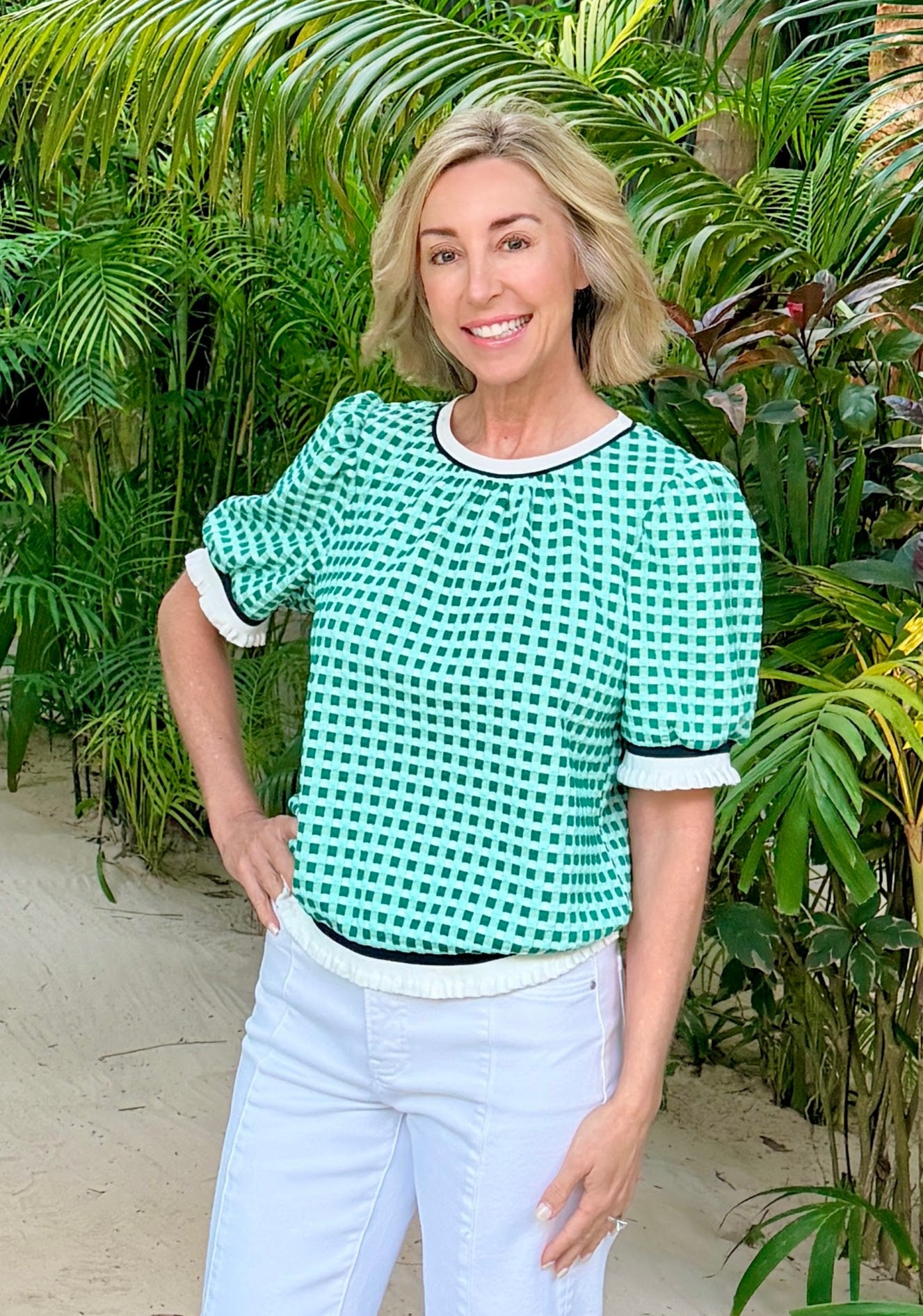 Woman wearing a green checkered top and light blue pants standing in front of green plants.
