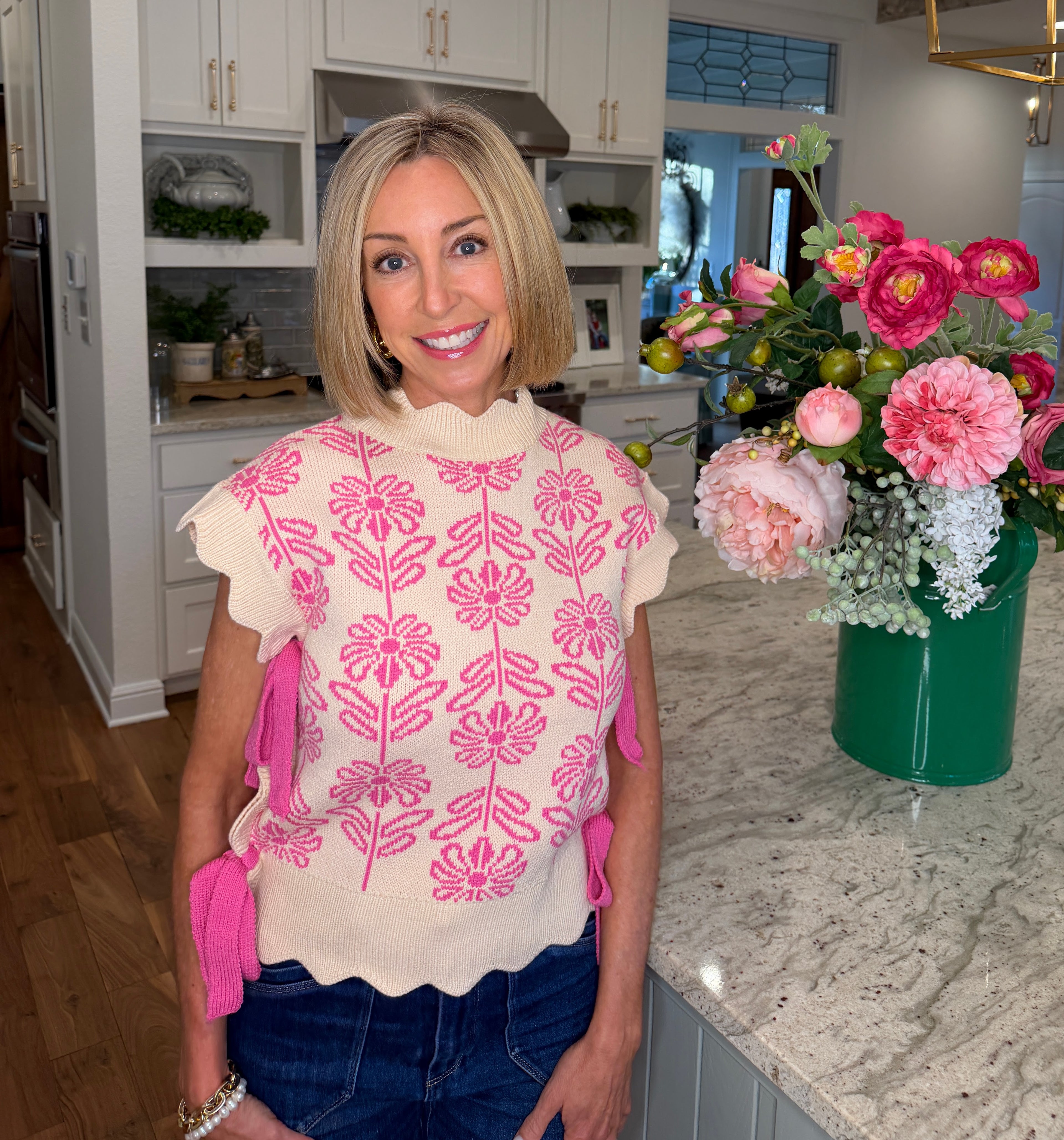 Woman standing in a kitchen with a floral-patterned top and pink gloves, next to a vase of flowers.