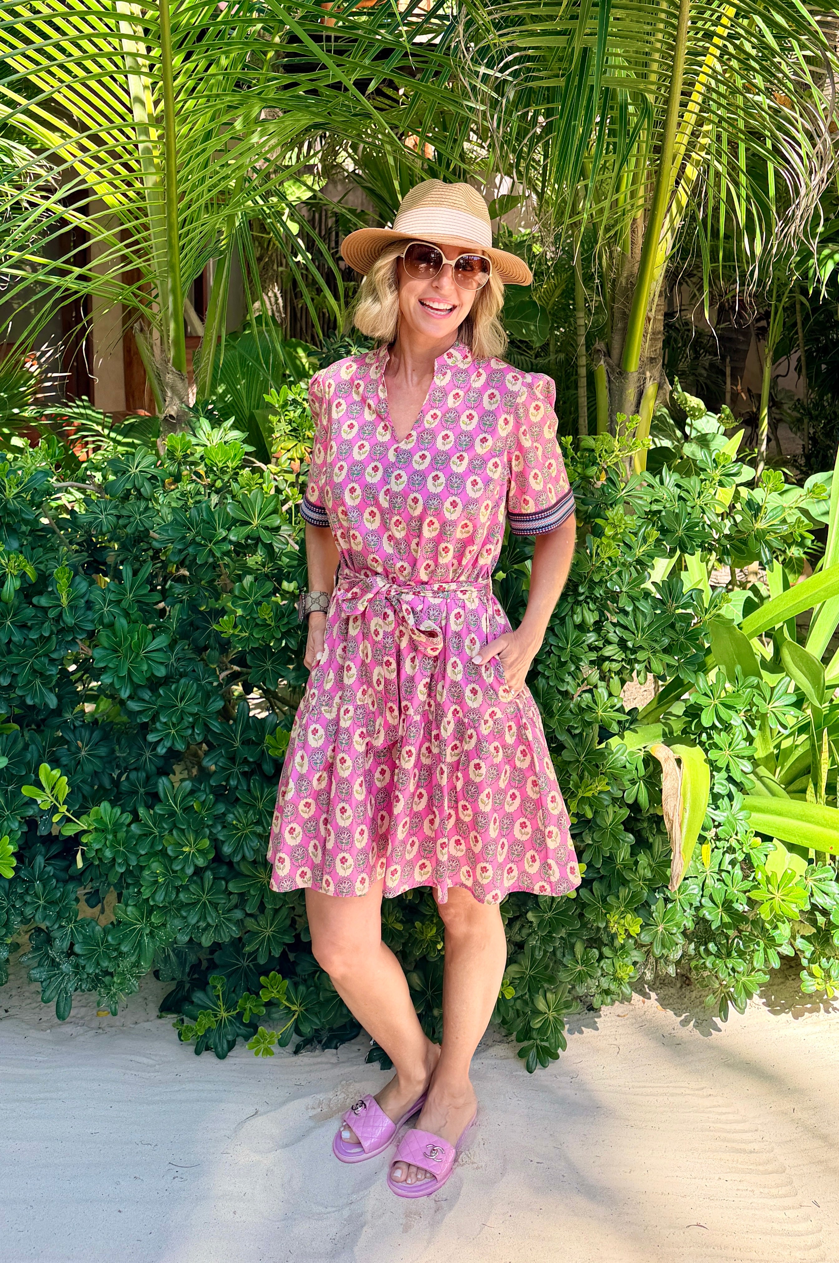 Woman in a pink patterned dress and hat standing in a tropical setting with palm trees.