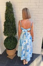 Woman in a blue and white patterned dress standing next to a potted plant against a brick wall.