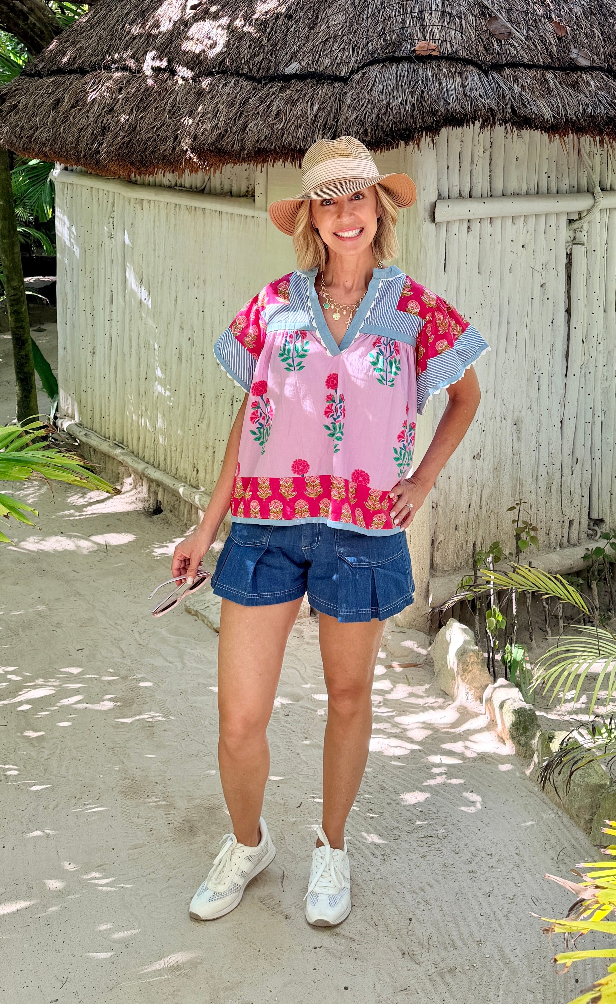 Woman in a colorful shirt and denim shorts standing in front of a thatched-roof hut.