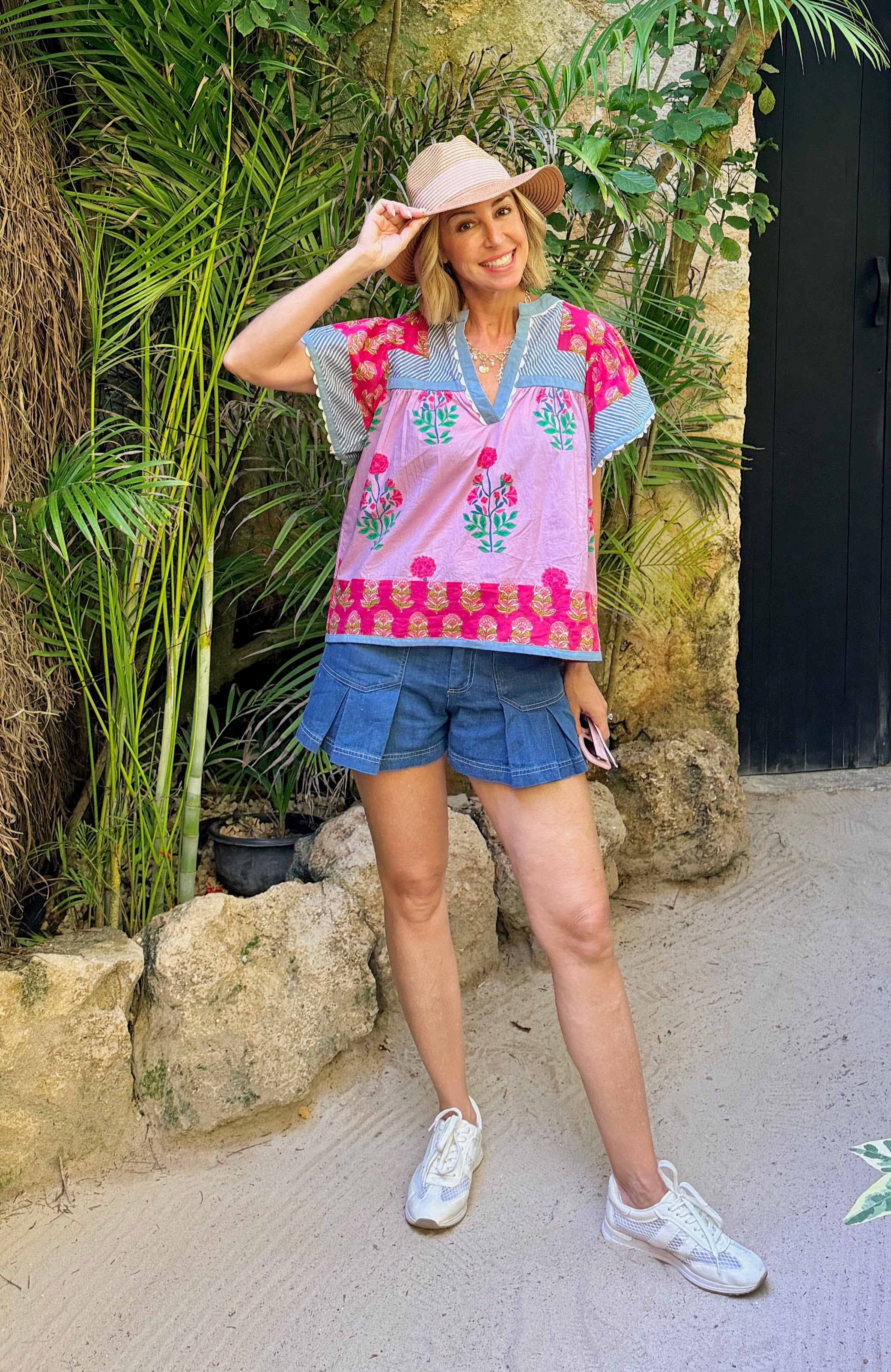 Woman wearing a colorful embroidered top and denim shorts standing in a tropical setting.