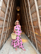Person in a pink dress with green patterns standing inside a thatched-roof structure.